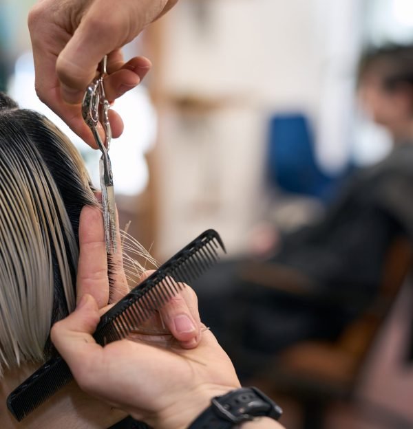 Hairdresser cutting hair tips of young female model with comb and scissors in hairdressing salon